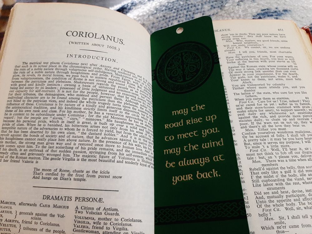 A photo of the title and introduction page of William Shakespeare's play "Coriolanus", with a green bookmark lying across the opposite page, obscuring one column of text; gold letters on the bookmark read: "May the Road rise up to meet you. May the wind be always at your back."
