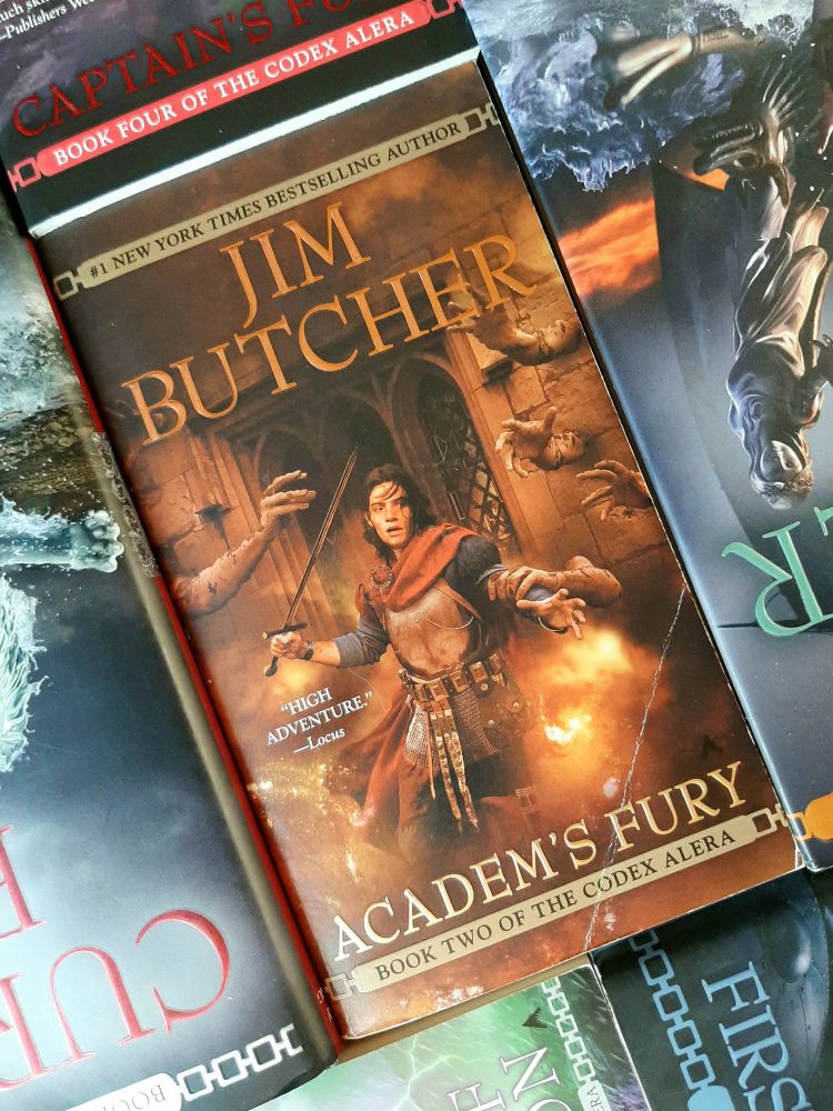 A photo of a somewhat creased paperback copy of Jim Butcher's second Codex Alera novel "Academ's Fury". Steve Stone's cover illustration depicts a dark-haired youth in Romanlike armor and scarlet cloak in the midst of a stone hall of reaching, grasping hands. Fire and smoke wreath the young man as he raises a long blade and recoils from the clutches of one of the hands.