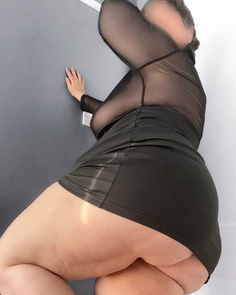 Camera looks up at Bianca who leans against the wall, showing off her round butt and thick thighs in a short black minidress that doesn’t fully cover her butt.