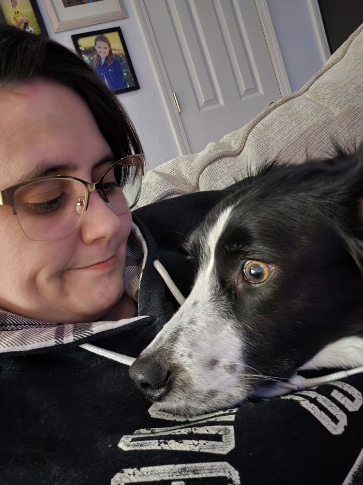 A woman with short dark hair and gold rimmed glasses sits on a beige couch with a black and white border collie rest her head on the woman's chest.
