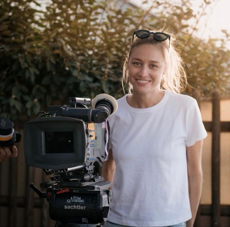 Woman posing with filming equipment and smiling, wearing white tee shirt and jeans