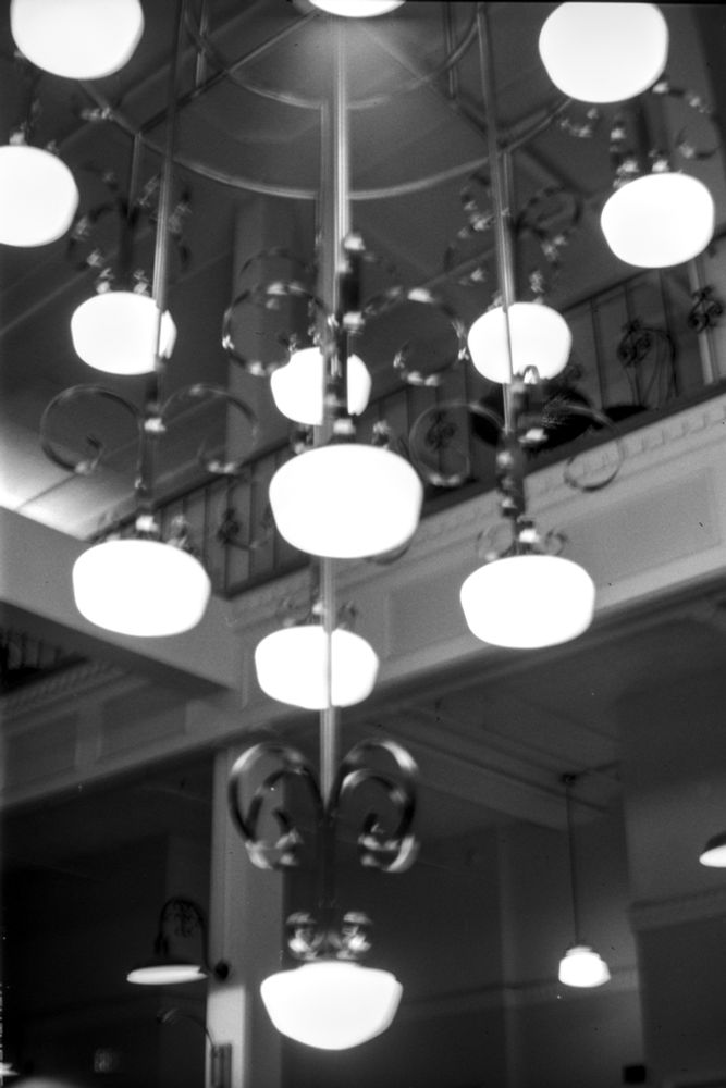 A black and white photo of a hanging light fixture with railings and higher floors above and behind it. 