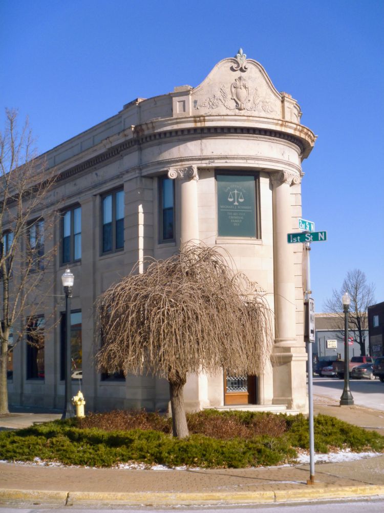 A white stone two-story building with a rounded front entrance against a clear blue sky. In front of the building is green landscaping and a leafless willow tree.  