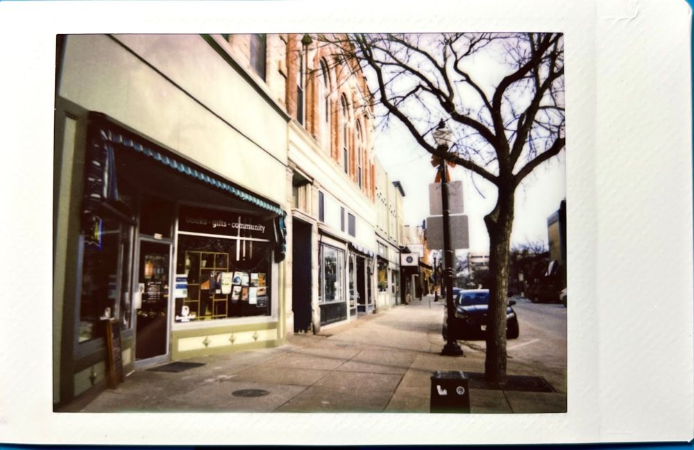 An instant photo showing several storefronts alongside a sidewalk with the sun directly hitting the buildings. 