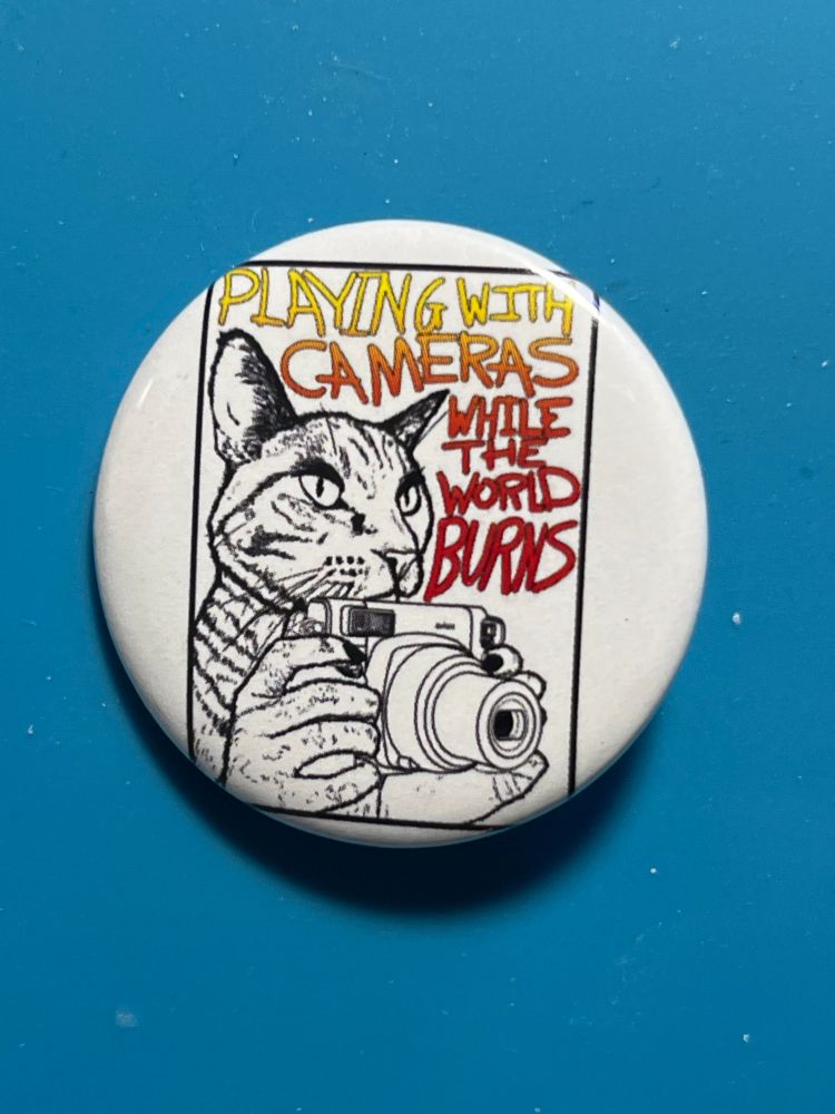 A photo of a button pin depicting a cat with hands holding a camera. Text behind the cat reads “Playing with cameras while the world burns” in a yellow-to-red gradient. 