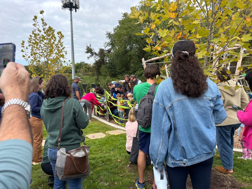 A crowd of people watching children and elected officials cutting green ribbons strung along a fence in front a natural looking climber and fall foliage visible around the play area.