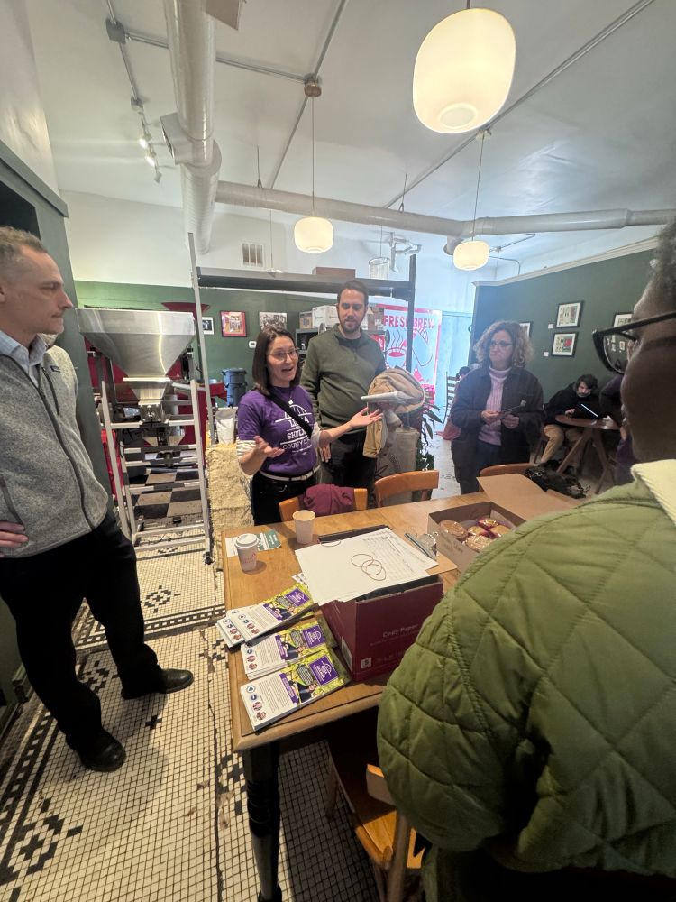 Lissa speaking to volunteers inside of a coffee shop. A table with Lissa literature is visible in the foreground.