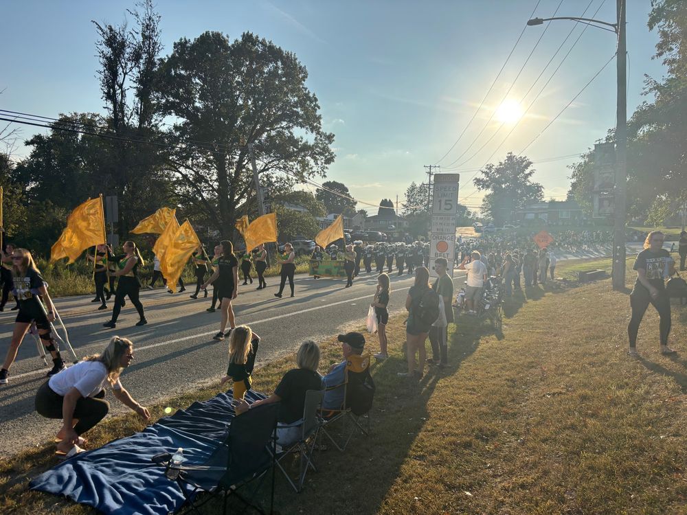 Deer Lakes marching band waving gold guard flags down a people-lined street with the sunsetting behind them.