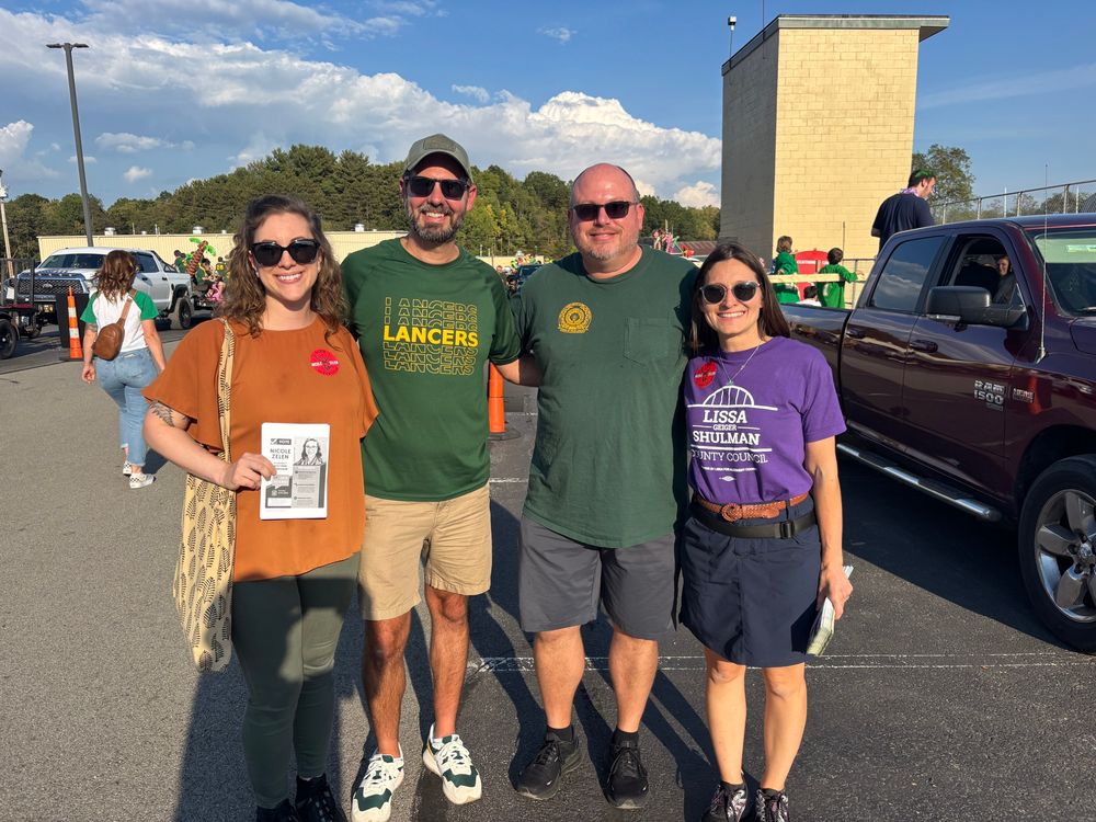 Lissa wearing a purple campaign T-shirt standing with two school directors in green Lancers shirts and candidate Nicole Zelen for Township Supervisor. Behind them parade floats are lined up in a school parking lot.