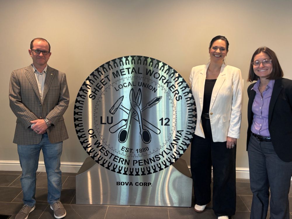 Lissa in a purple button down blouse and blazer standing next to a woman in a white blazer and a metal statue of the Sheet Metal workers logo. A man in jeans and a blazer is pictured on the other side of the metal logo.