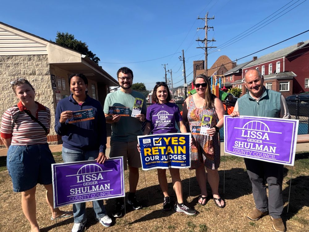 Group of people smiling at camera with Lissa for PA purple lawn signs, a Vote YES to retain our judges lawn sign, campaign literature, and a playground visible behind them.
