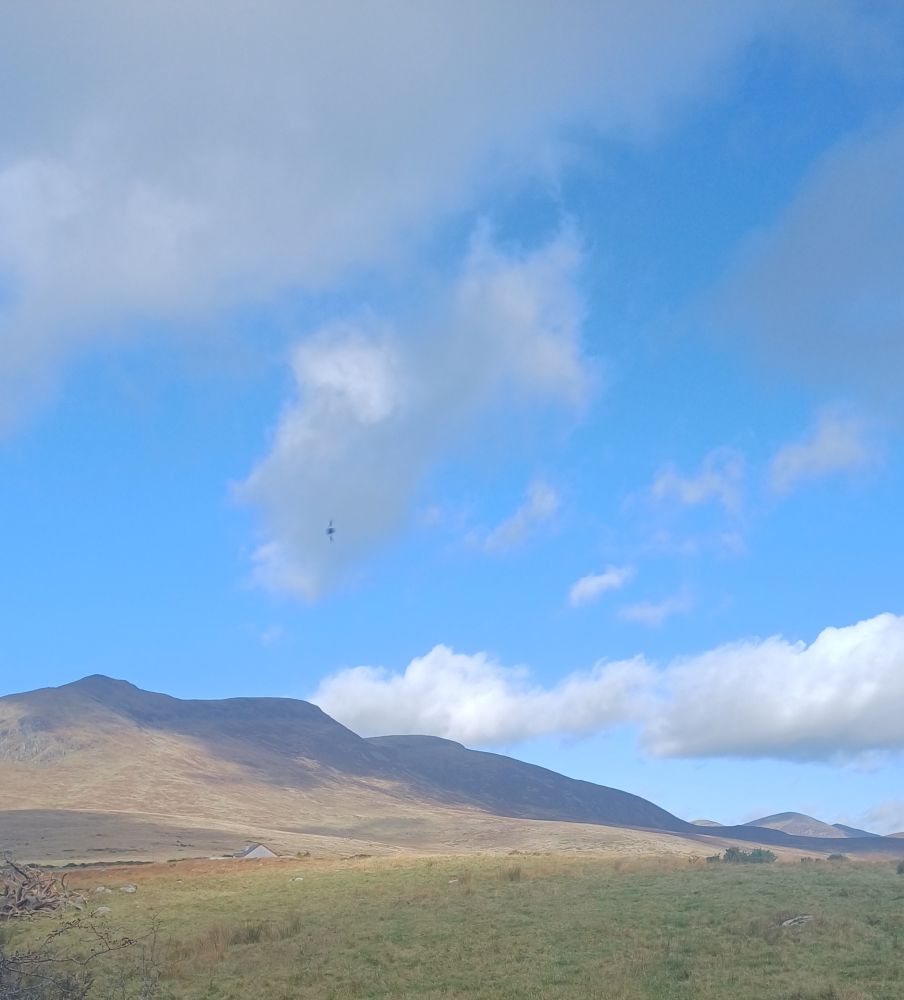 The mountains of Mourne in an autumnal light. There are cloud shadows across the mountains. The sky is big and a light aqua blue.