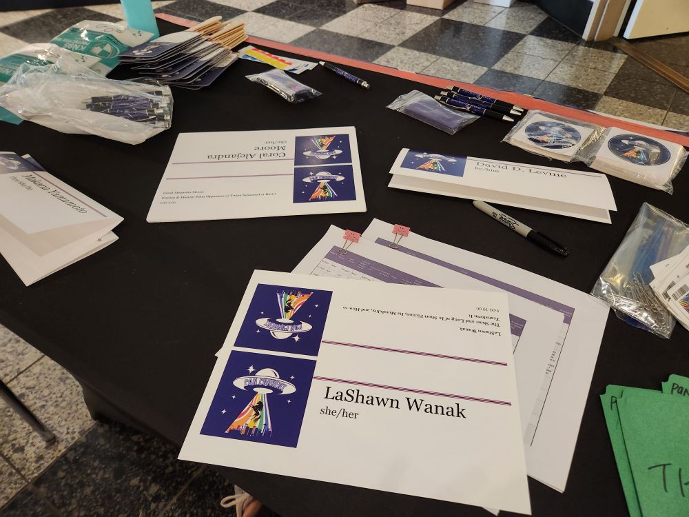 Table tents with panelist names are spread across a table with a black tablecloth. ConCurrent pens, stickers, fans, and badge ribbons are visible in the back of the photo.