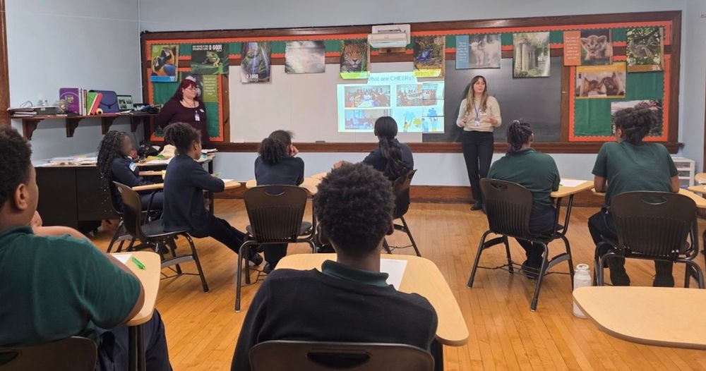 Classroom scene with students in uniforms listening to a person presenting at the front near a chalkboard with educational posters.