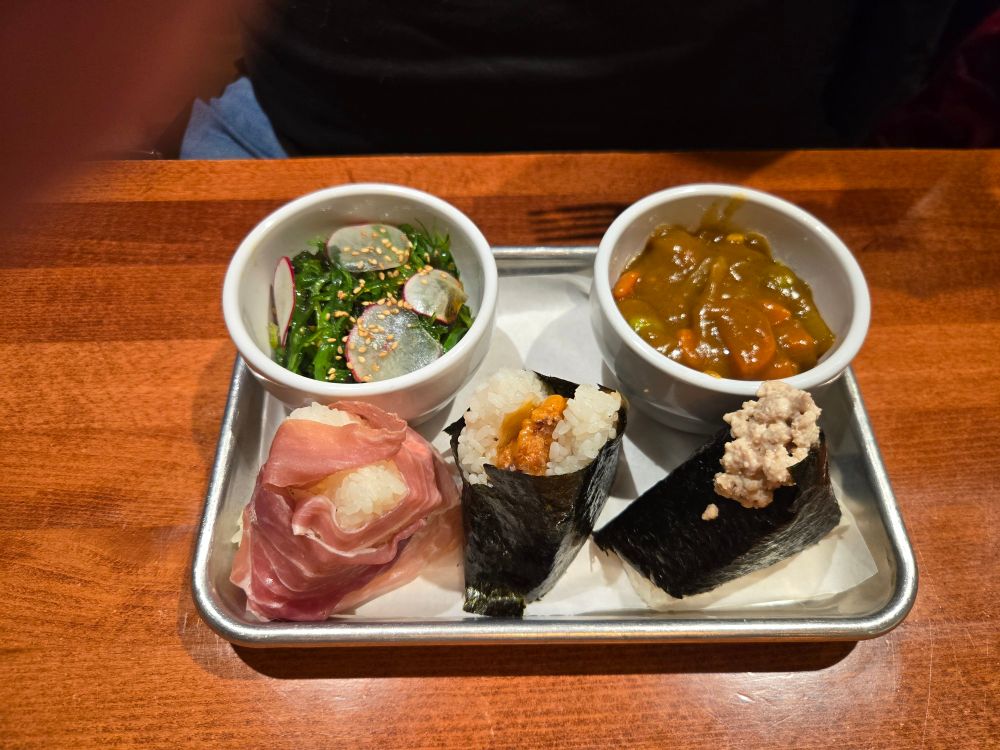 Three onigiri (one wrapped in prosciutto instead of seaweed) on a tray with bowls of cucumber salad and vegetable curry.