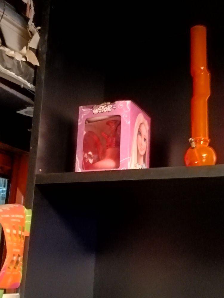 Caixa contendo um narguilé rosa da Barbie em uma prateleira preta ao lado de um bong de cano alto 