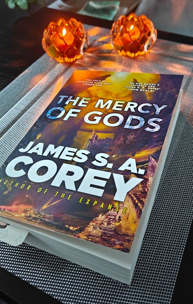 A paperback copy of the novel The Mercy of Gods by James S.A. placed on a table next to two beautiful candles.