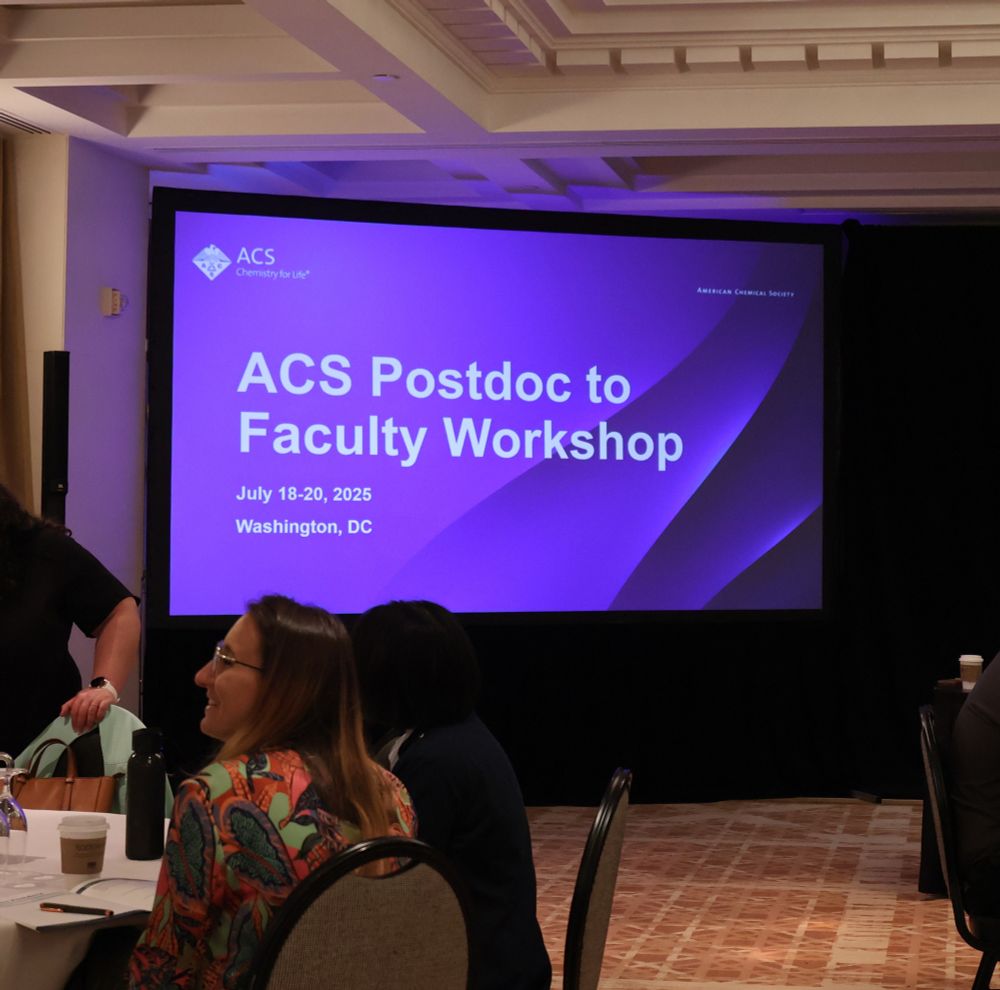 Room with round table of people sitting infront of a screen projecting the text ACS Postdoc to Faculty Workshop with dates and location. 