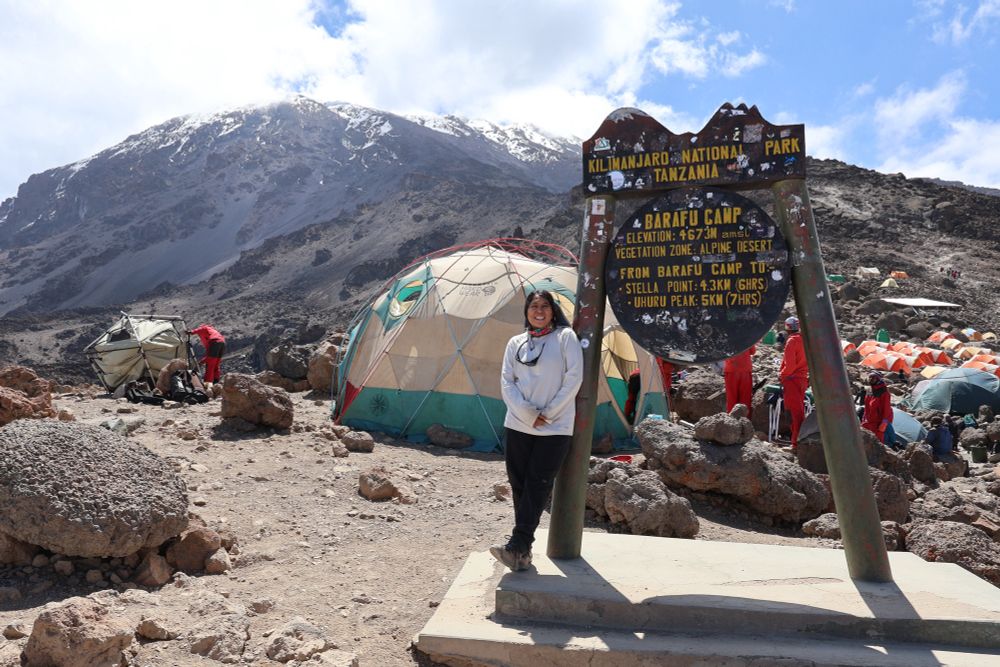 Person leaning on a wooden sign with text: Kilimanjaro National Park Tanzania Barafu Camp. In the background is the peak of Mt. Kilimanjaro. There are also tents and people at camp in the background. 