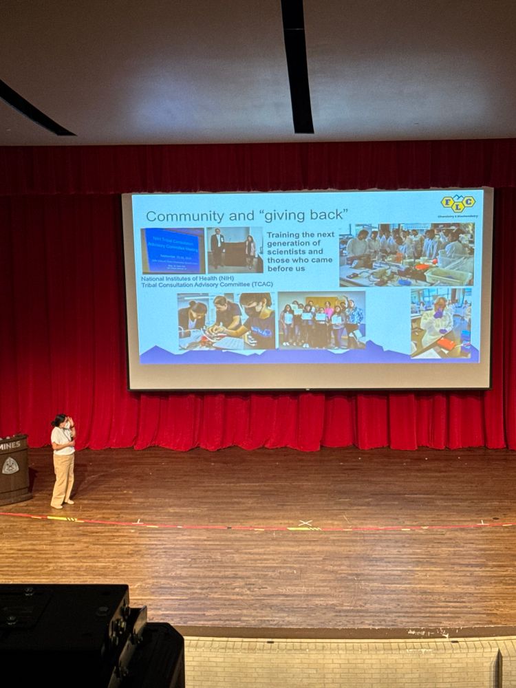 Woman standing on stage looking towards the screen projecting photos under the text “community and giving back”. 