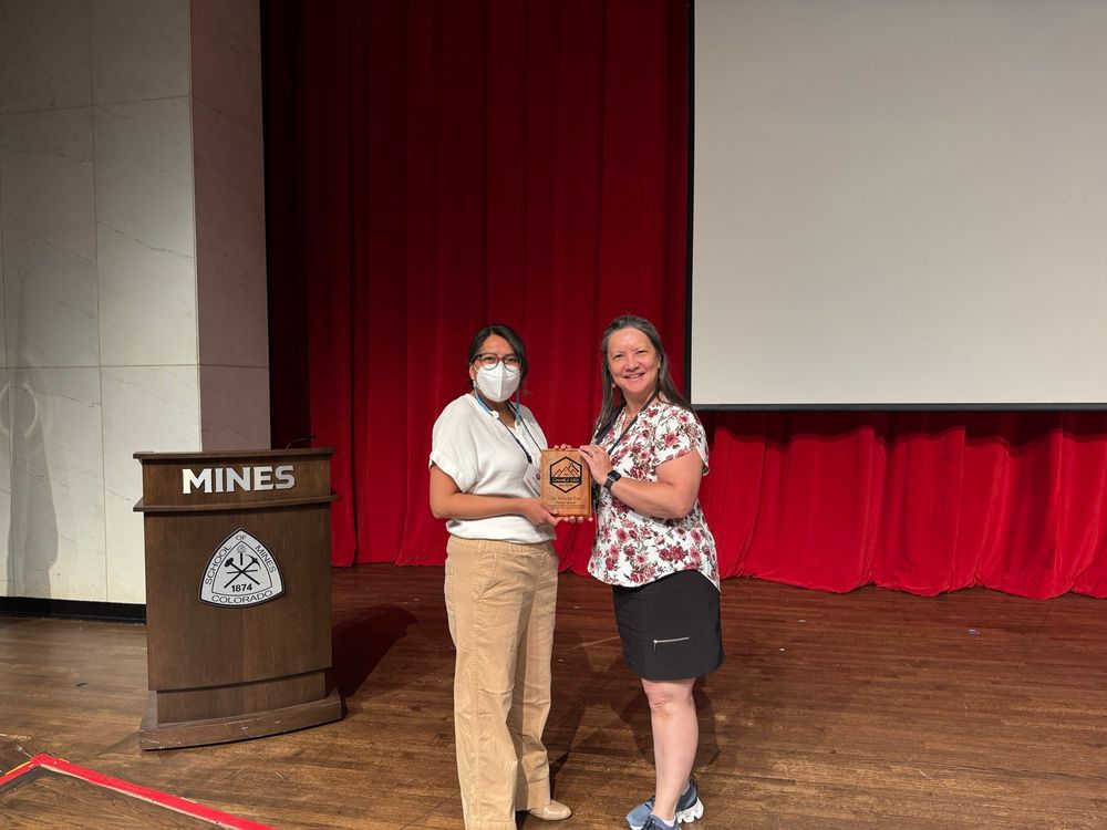 One woman masked holding a wooden plaque next to another woman, both looking at the camera. In the background is a podium with Mines and its emblem. 