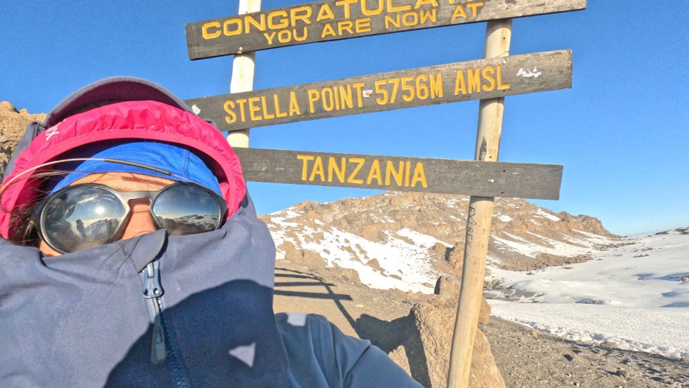 Person wearing sunglasses and zipped up jacket in-front of sign that reads Congratulations you are now at Stella Point 5756 m Tanzania. In the background is the crater of Mt. Kilimanjaro covered with snow and ice. 