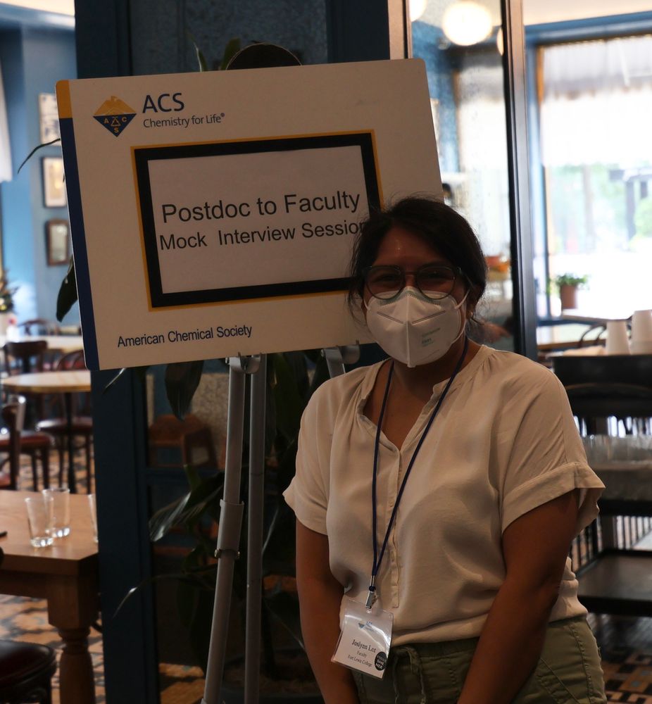 Masked person wearing a badge standing infront of a sign that reads Postdoc to Faculty Mock Interview Session. 