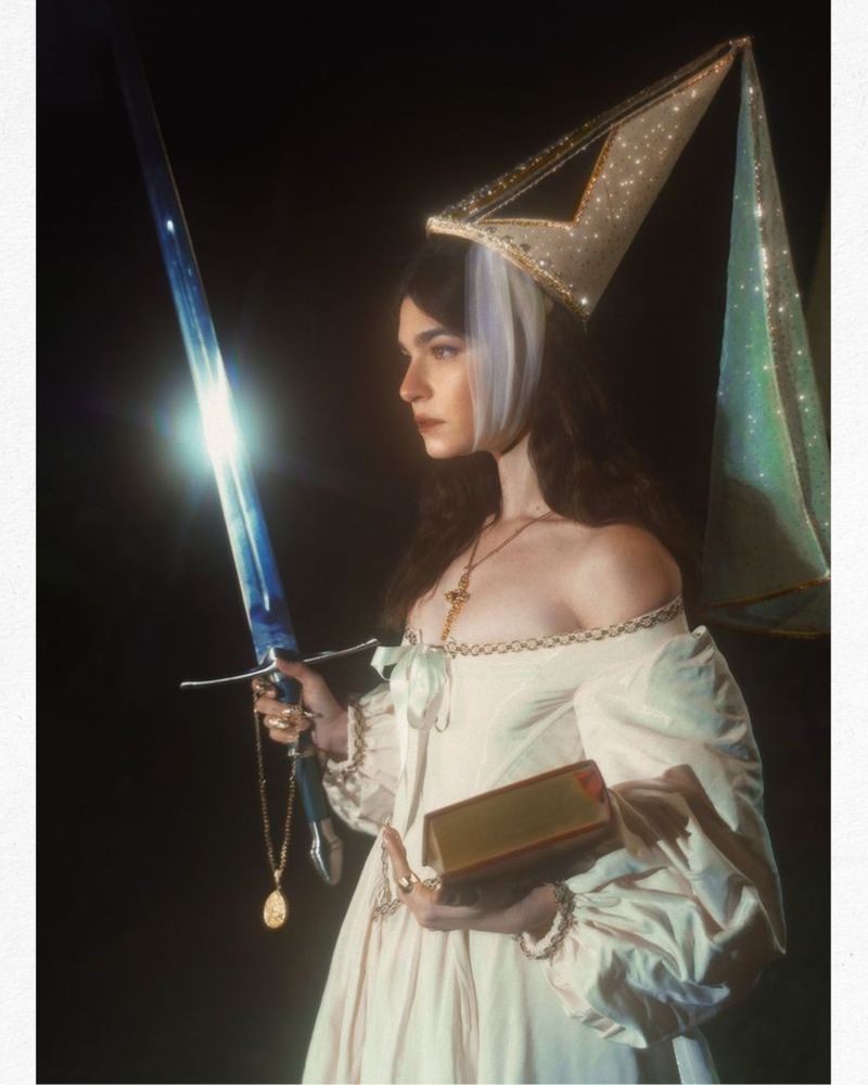 The reference photo I used for my piece! This is a photoshoot of a person dressed up as a 15th century princess, done by Emackphoto on Instagram. This person holds a steel sword in their right hand and a book in their left. They wear a white, off-the shoulder, medieval dress and a sparkling hennin on their head. The background is completely black. 