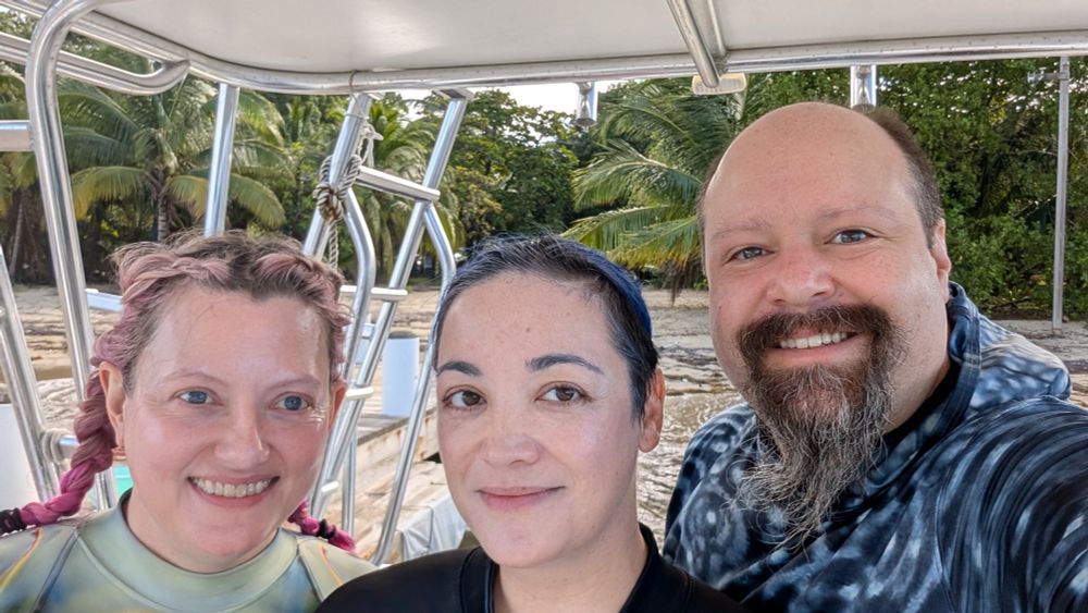 A close up photo of a smiling group of three people: one with pink braids, one with blue hair pulled back, and one with a goatee. They are standing on a boat at shore 