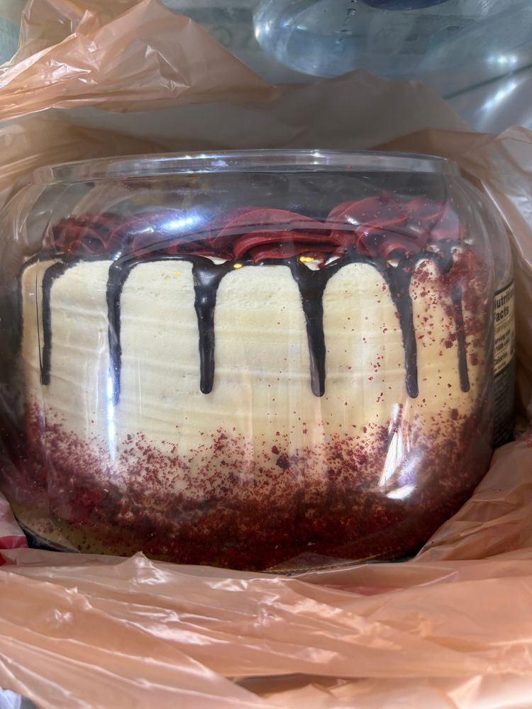 A cake with white frosting, red decorations and chocolate draped down the sides in a cake container.
