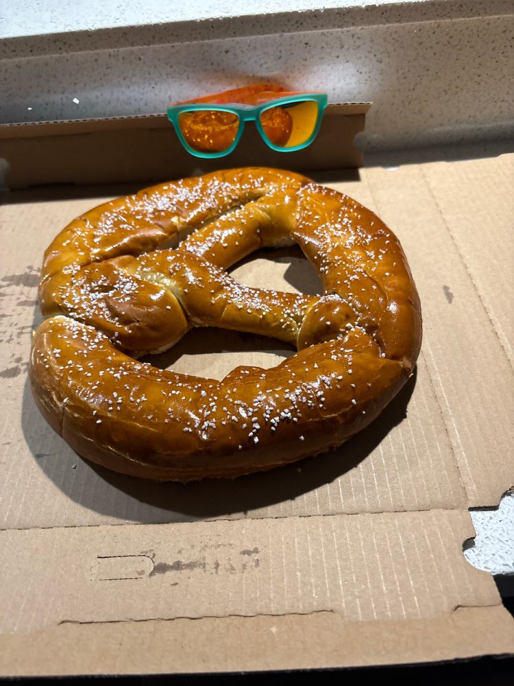 A very large soft pretzel in a pizza box with a pair of mirrored sunglasses on the side of the box. The pretzel is about 3 times as wide as the sunglasses.