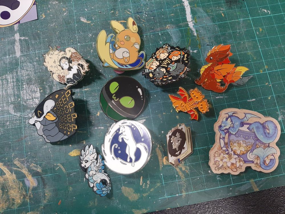 A bunch of enamel pins and one wooden pin, waiting to be arranged in the snita bag