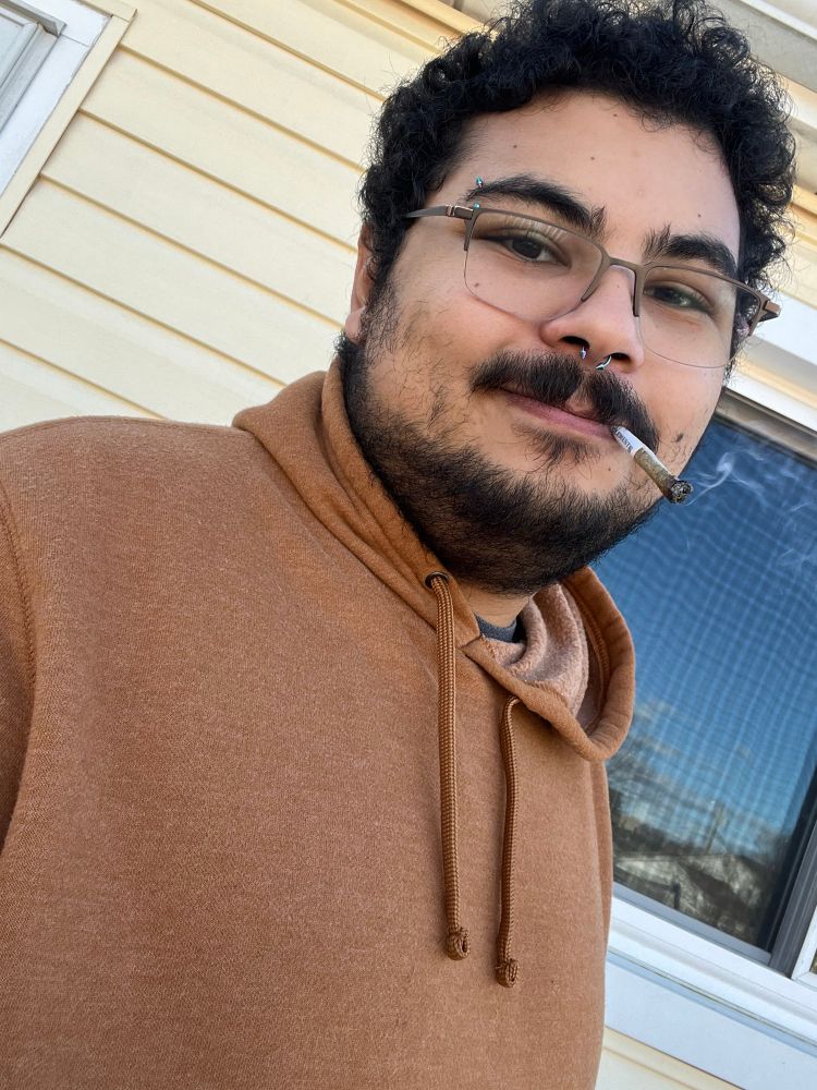 Teddy stands outside in a brown sweater, smiling slightly and smoking a joint.