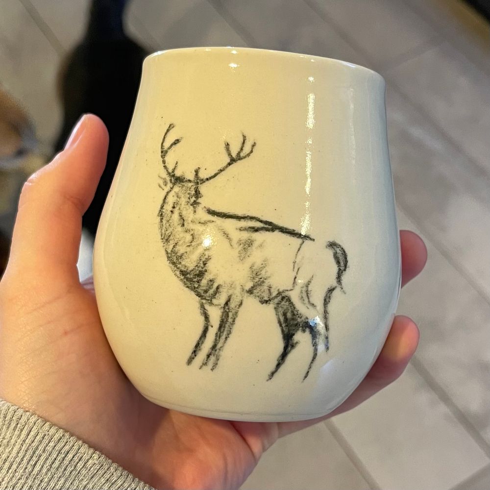 A hand holding a white ceramic cup with a sketch of a stag on it. The cup is the shape of a stemless wine glass and is covered in a glossy clear glaze. 