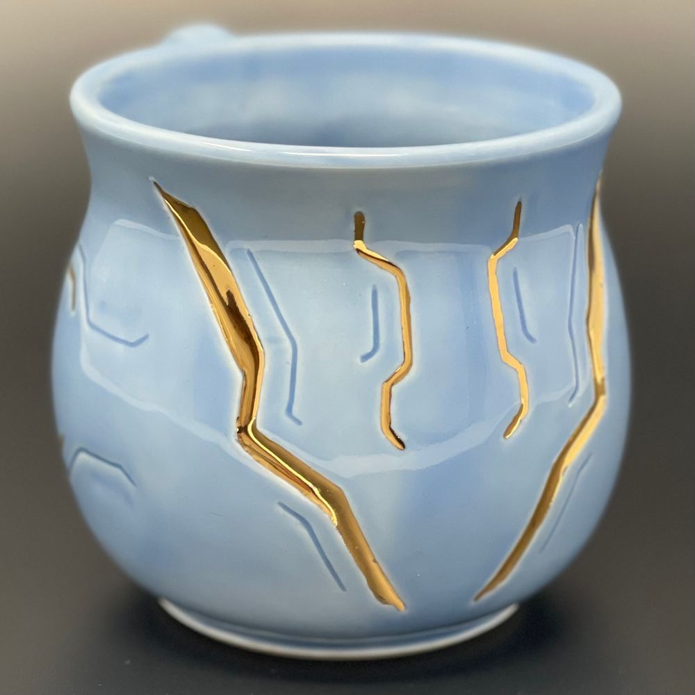 a close-up photograph of a ceramic mug with a sky blue-colored glaze. the mug is carved to resemble cybernetic implants from the video game Cyberpunk 2077, and some of the carvings have been filled in with gold luster.