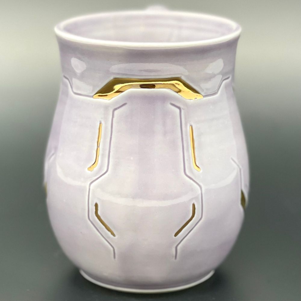 a close-up photograph of a ceramic mug with a lavender-colored glaze. the mug is carved to resemble cybernetic implants from the video game Cyberpunk 2077, and some of the carvings have been filled in with gold luster.