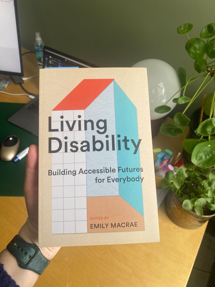 A photo of a book called Living Disability by Emily Macrae. The book is being held over a desk with a plant and some pens in the back.