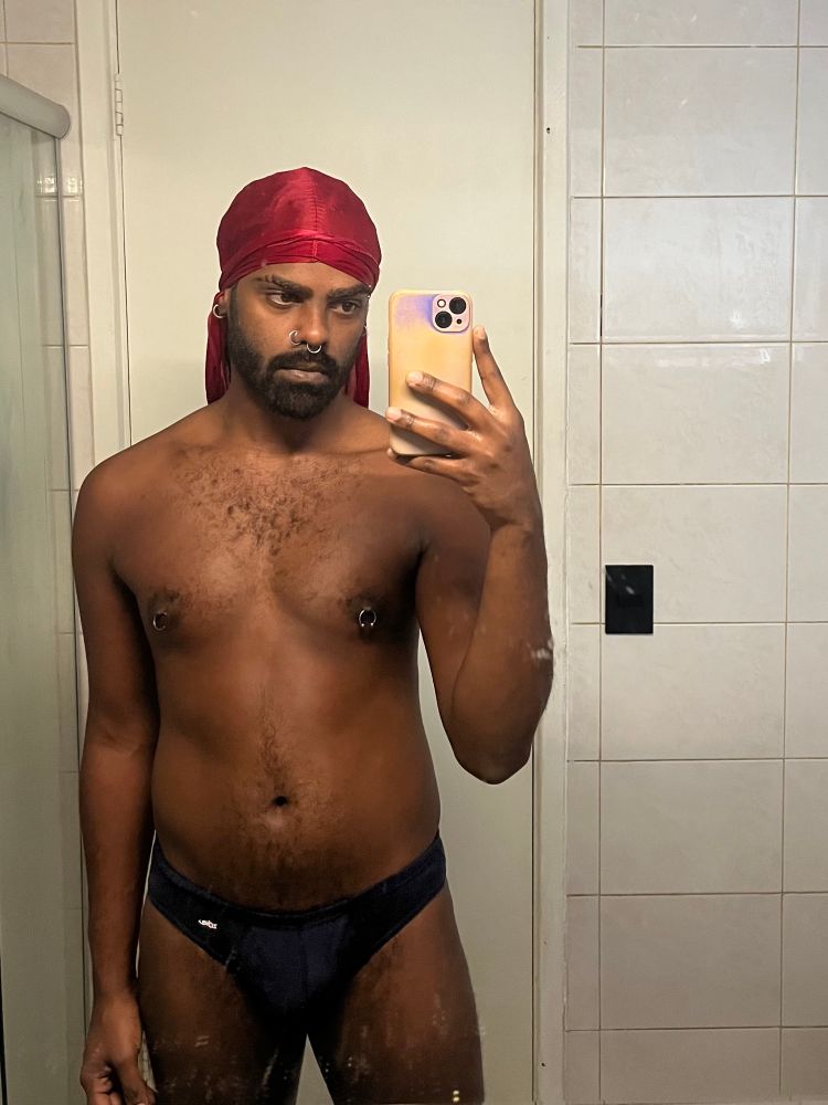 eu sou um homem preto de durag rosa piercing nos mamilos tirando uma selfie segurando o celular na mão direita em frente ao espelho 