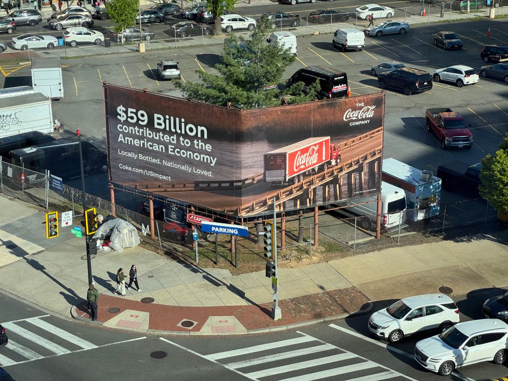 Billboard for Coca-Cola reading “$59 billion contributed to the American economy” showing a picture of a Coca-Cola truck driving across the bridge. 