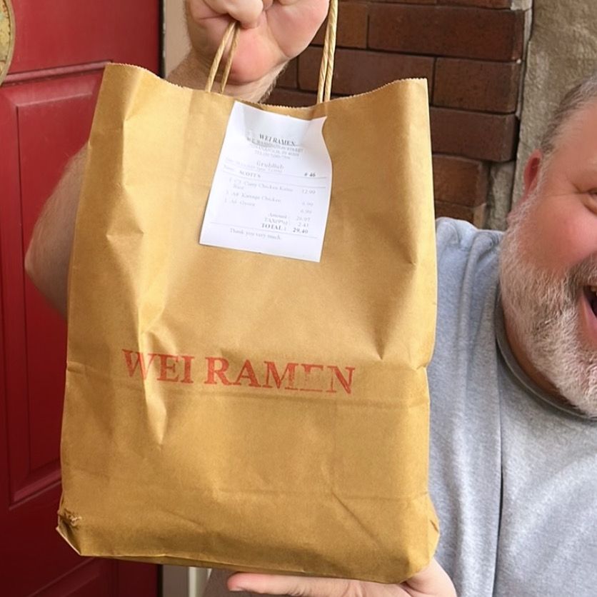 A bearded man (of whose cheesily-grinning face we can only see about a third) holds a to-go bag from Wei Ramen in front of a red door and brown brick wall