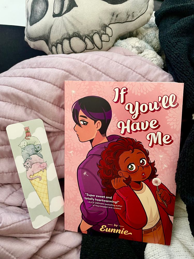 An image of a book leading against a pile of blankets. The book is titled “If you’ll have me” by Eunnie. On the cover a tall tan skinned person with short, straight black hair stands facing bf away from the viewer, their face turned to look down and back at the character in the center. The central character is posed facing the reader with her back to the other protagonist so their backs are touching. The shorter feminine character has warm dark skin, glasses, and auburn wavy hair. She holds a dandelion, as if in reference to making a wish.
 Above the book half of a skull looking pillow is visible. To the left of the book is a bookmark that depicts an ice cream cone where instead of ice cream cats of various colors are piled on top. 
