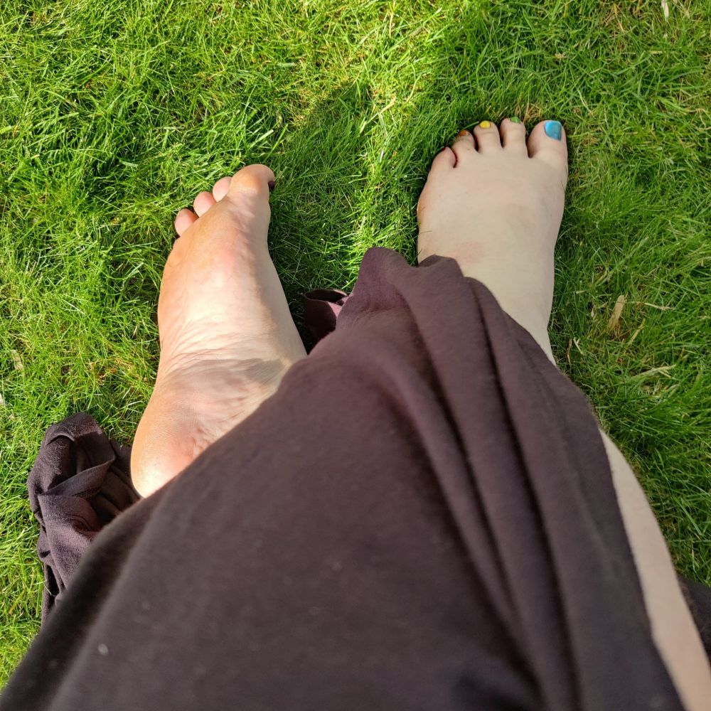 Der Blick von oben auf meine überschlagenen Beine auf grüner Rasenfläche. Die Füße und ein Teil eines Schienbeins schauen unter meinem langen, schwarzen Rock heraus. Die Zehennägel sind in Regenbogenfarben lackiert. 