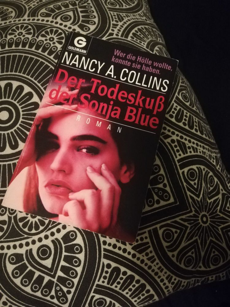 Auf einem schwarz weiß gemusterten Kissen liegt ein Buch mit dem Titel "Der Todeskuss der Sonja Blue". 