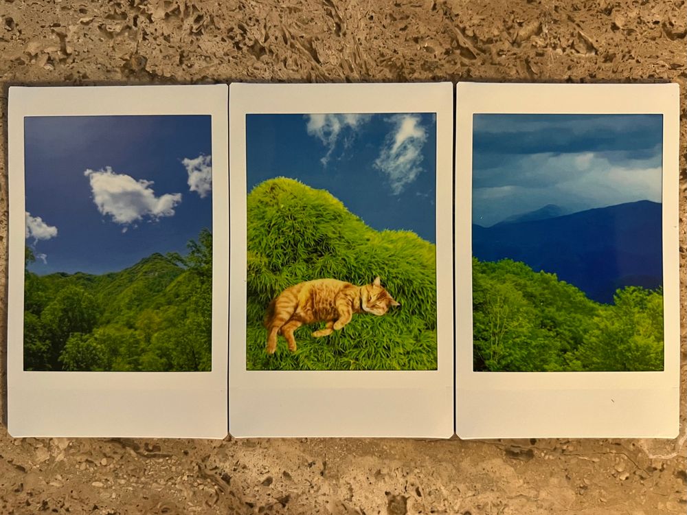 A photograph of three Instax Mini photographs set side by side on a marble table. On the left is a photo I took in the Smoky Mountains, with a bold blue sky, bright green leafy tree-covered mountain ridge, and a few cotton candy clouds. On the right is a photo Jonathan took in the Smoky Mountains, with a bright green treeline in the foreground, moody blue mountains in the background, and a cloudy sky with layered veils of blue. In the center is a photo collage, with a bright blue sky and some wispy white clouds, and a bright green mound of fuzzy textured moss, with an orange tabby cat superimposed, looking like he’s asleep on his side, nestled into the moss. The way the photos are lined up, there seems to be one curving horizon of green against blue that continues through all three images.