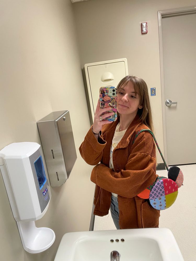 mirror selfie of a short girl w brown hair smiling wearing a rust colored jacket, an off white graphic tee& jeans carrying a colorful mickey mouse shaped purse 