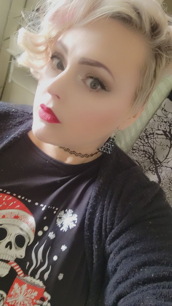 Blonde woman in bright red lipstick with gothic Christmas accessories and a shirt with a skeleton in a Santa hat drinking hot chocolate. 