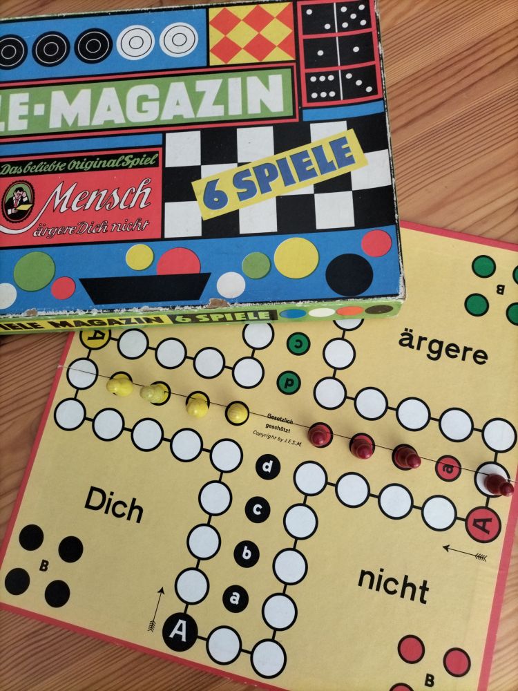 Mensch-ärgere-dich-nicht-Spielfeld bei dem Rot die letzte Figur direkt vorm Haus hat und Gelb gewonnen. Darüber der Karton der Spielesammlung für 6 Spiele. Leider fehlt der Würfel mit der 6 nach oben. Aber wer will schon perfekt sein. 😂