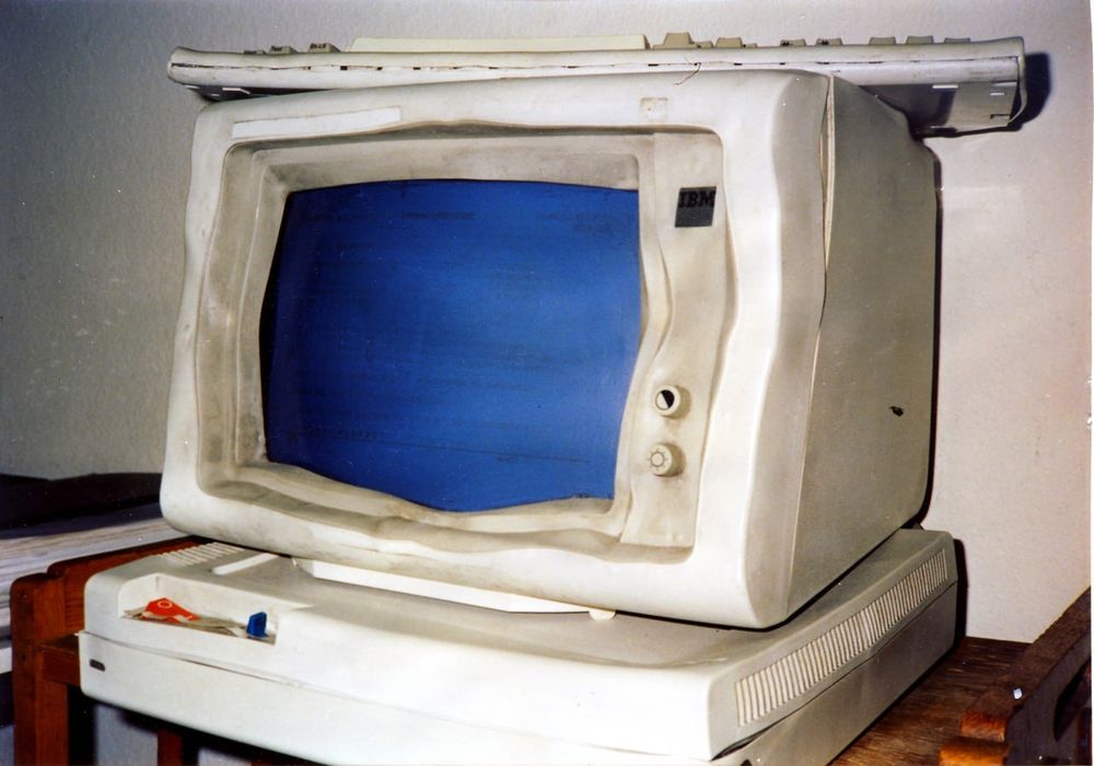 Ein verformter Computer-Monitor steht auf einer horizontalen, ebenfalls etwas ramponierten Rechner-Einheit. Oben drauf liegt eine Tastatur.