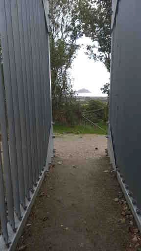 A narrow path between two grey metal fences, leading towards a background of grass and shrubs. Beyond them is the sea, with a small, slightly hazy island in the distance. The pale grey of the sea is indistinguishable from the sky.