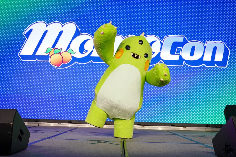 Mo, a large green three-eyed monster, dances in front of a blue background and the MomoCon logo.